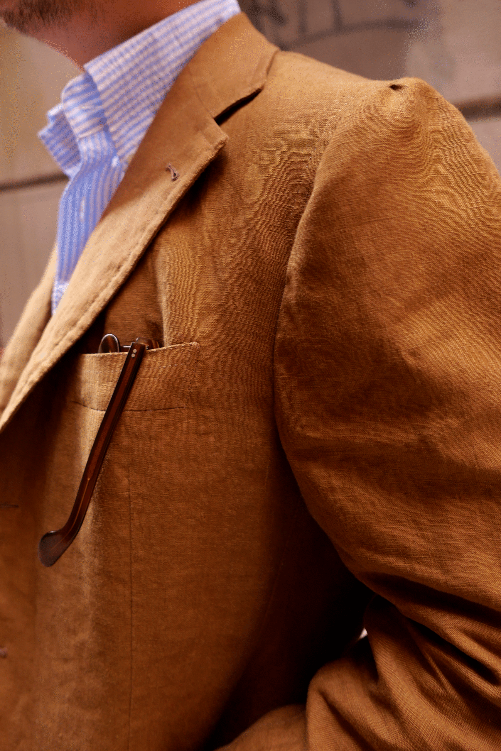 Close-up of a Neapolitan linen jacket in warm brown, softly structured with natural shoulder and visible texture, paired with a light blue striped shirt and glasses in the breast pocket, expressing relaxed summer elegance.
