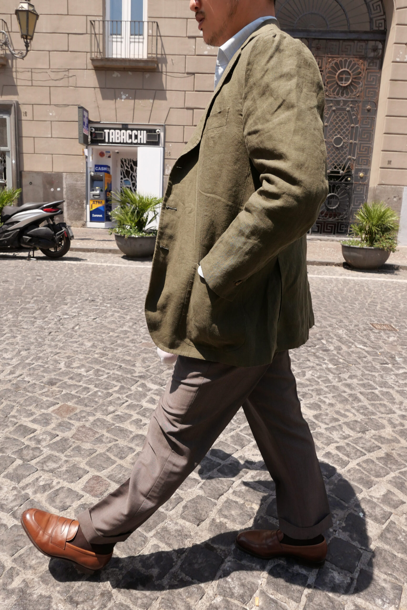 A man wearing bespoke Neapolitan tailoring—an olive linen jacket with taupe trousers and brown loafers—walks along a sunlit cobblestone street in Naples.